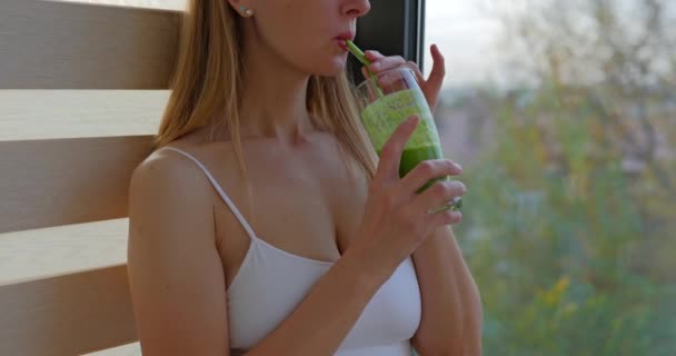 スポーティな女性は緑のスムージーのガラスと窓の近くに滞在しています ストローから緑色のスムージーを飲む少女 高品質の4K映像 — ストック動画