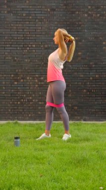 Antrenmandan önce saçını bağlayan bir kadın, tuğla duvarın yanında çimlerin üzerinde duruyor. Yüksek kalite 4k görüntü