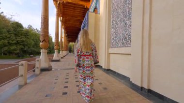 Özbekistan 'ın Taşkent kentinde düzenlenen Anma Töreni' nin önünden geçmekte olan geleneksel Özbek süsleme desenli kıyafetli bir kadın görülüyor. Mimaride oyulmuş ahşap sütunlar ve Özbek mirasını yansıtan mavi bir giriş yer alıyor..