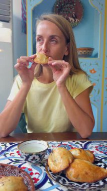 Geleneksel bir Özbek kafesinde oturan bir kadın taze pişmiş samsa hamur işi yiyor. Masa ulusal seramik yemeklerle süslenmiş, Özbek mutfağını, kültürünü ve yerel yiyeceklerin tadına bakmanın keyfini yansıtan otantik bir atmosfer yaratıyor..