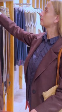 Kahverengi bir ceket ve bej bir çanta giymiş bir kadın modern bir giyim mağazasında deri kemerleri dikkatle inceliyor. Sahne, çağdaş alışveriş, perakende moda, tüketici seçimi ve kentsel bir butik iç mekanının şık atmosferini yakalıyor..