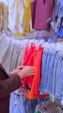 Modern bir giyim mağazasında kadın eli, canlı bluzları ve kot şortları inceliyor. Görüntü, perakende ortamda modaya uygun kıyafet alışverişi, tüketici yaşam tarzı, mevsimlik moda ve şık tercihleri vurguluyor.