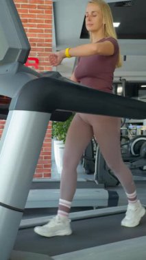 Modern bir spor salonundaki koşu bandında yürüyen bir kadın antrenmanına odaklanıyor. Bileğine spor aleti takıyor, aktif yaşam tarzını, sağlığını ve fiziksel sağlığını vurguluyor. Görüntüler spor motivasyonu ve kardiyo eğitimini temsil ediyor..