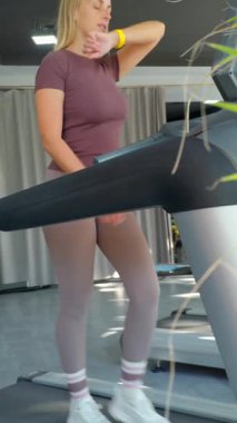 Spor kıyafetli genç bir kadın koşu bandında yürüyor ve aktif bir egzersizden sonra yüzünden ter siliyor. Spor salonu parlak ve modern, sağlıklı bir yaşam tarzı, günlük egzersiz ve düzenli fitness eğitimi yoluyla vücut bakımı motivasyonunu vurguluyor..