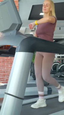 Spor giyimli genç bir kadın koşu bandında antrenman yapıyor ve kalp atışlarını akıllı yama kullanarak kontrol ediyor. Modern spor salonu, aktif bir yaşam tarzının sürdürülmesi için günlük çalışmalara uygunluk, sağlık izleme ve teknoloji entegrasyonunu yansıtır..