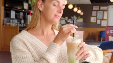 Beyaz örgü kazaklı bir kadın, rahat bir kafedeki ahşap bir masada oturur ve pipetle buzlu kibritli latte içer. Gözlerini kapatıyor, rahatlatıcı zevkin ve rahatlatıcı anın tadını çıkarıyor, sıcak iç aydınlatma ve sakin ortam ile çevrili..