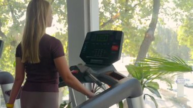 Spor giyimli genç bir kadın, doğal ışık ve yeşillikle dolu modern bir spor salonunda koşu bandında yürüyor. Büyük pencereler, sabah antrenmanı için sakin ve ilham verici bir atmosfer yaratıyor. Aktif bir yaşam tarzı için sağlık, denge ve motivasyonu teşvik ediyor..