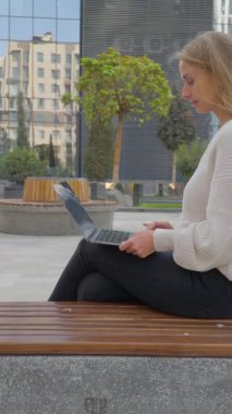 Özbekistan 'ın Taşkent kentindeki modern bilişim parkı binalarının yanındaki bankta otururken, beyaz kazaklı sarışın bir kadın laptopuyla çalışıyor. Görüntü uzaktan çalışmayı, dijital yaşam tarzını ve bir teknoloji merkezinin modern kentsel ortamını temsil eder..