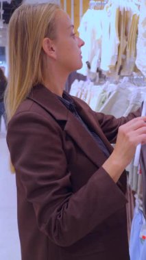 Moda mağazasından gri kolsuz bluz seçen bir kadının yakın çekimi. Görüntü, modern bir perakende pazarlama ortamında modaya uygun kıyafetler, mevsimlik koleksiyonlar, şık kıyafet seçimleri ve tüketici yaşam tarzını ön plana çıkarıyor..