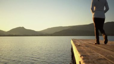 Modaya uygun şapkalı genç bir kadın, yürür ve sonra durur ve bir gölün ya da okyanusun ahşap rıhtımında geri döner, doğanın güzelliğine ve dağların arasında güneşin batışına hayran kalır.