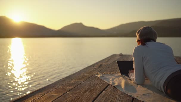 自由职业选择的概念 一个年轻的女人在她的笔记本电脑上工作 在日落时躺在湖畔或码头上 在美丽的自然环境中 通过在大自然中生活来改善生活质量 — 图库视频影像
