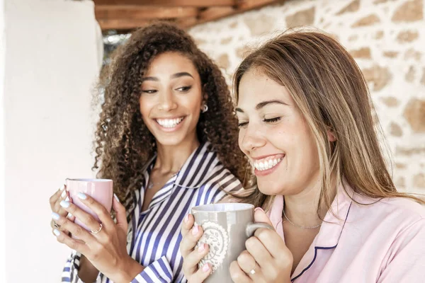 New family habits: multiracial female couple they drink a cup of tea on the porch of their country house - Two beautiful young women drinking coffee and smiling