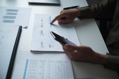 Profesyonel iş adamı, girişimci ya da muhasebeci masasında cep telefonu kullanarak mali rapor evrakları üzerinde çalışıyor..