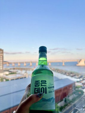 Busan, South Korea - 11 Aug 2024: Holding Soju in one hand with background as Gwangandaegyo Bridge aka Diamond Bridge a suspension bridge located in Busan, South Korea at Golden hour. clipart