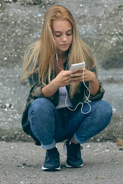 Squatting Flicka Absorberas Att Titta Smartphone När Bär Hörlurar Urban — Stockfoto