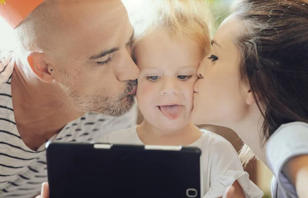 Close-up picture of two parents kissing their little girl on her cheeks while she plays with the tablet (she stucks her tongue out)