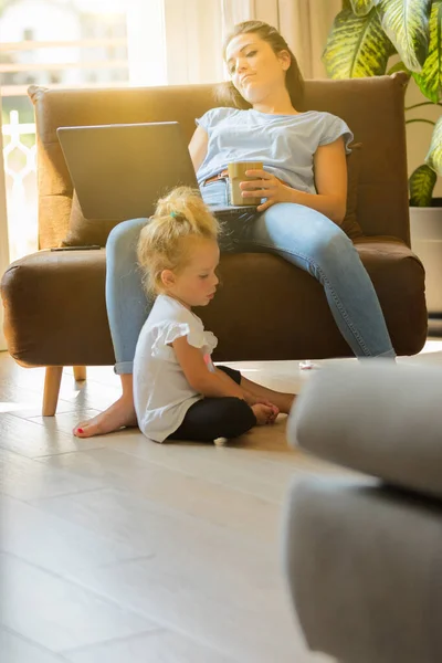 肘掛け椅子に座った母親は 大きなカップから飲み物を飲みながらノートパソコンを使い 子供は足で床で遊んでいます 家のリビングルームで暑い夏の日 — ストック写真