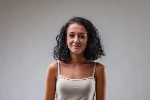Frontal portrait of a young brunette girl in a tank top; she has a controlled, calm, perhaps restrained smile; she is tanned and wearing a tank top