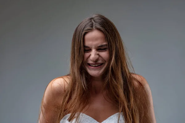 Frontal portrait of a young brown-haired girl wearing a white sheath dress laughing or smiling, unable to restrain herself from bursting into laughter