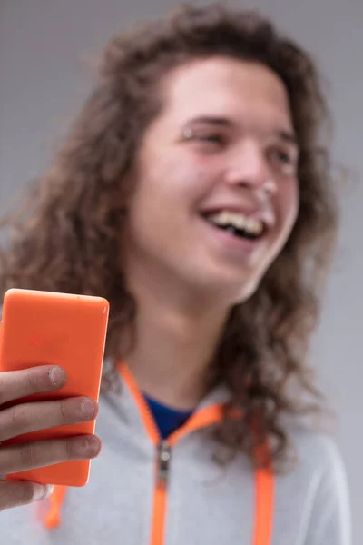 头戴长帽 眉毛刺痛的年轻人 背景模糊了 在前景的焦点是他的手拿着一个橙色的智能手机 他笑着 穿着运动衫 — 图库照片