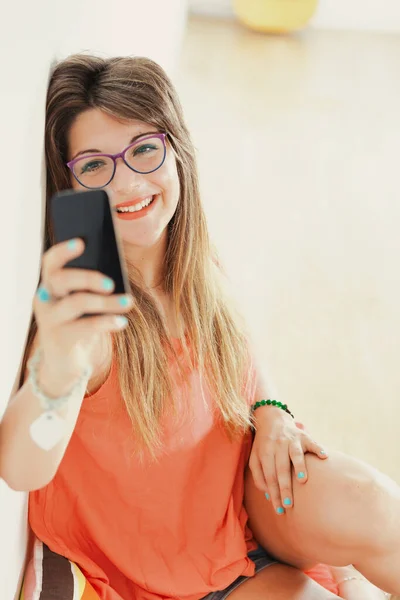 眼鏡を背負ったまま家の壁に寄りかかっている女性を応援しながら 自撮りやビデオ通話をするスマートフォンを持っている — ストック写真