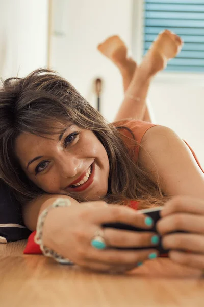 女人躺在地板上 手里拿着智能手机看视频或发短信 她有一头棕色的长发 微笑着看着摄像机 基层框架 — 图库照片
