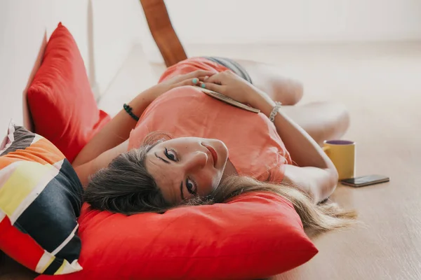 Young woman lying on her back on the floor between soft pillows with a notebook on her belly. She has long brown hair and looks into the room with serious curiosity. Different Tint.