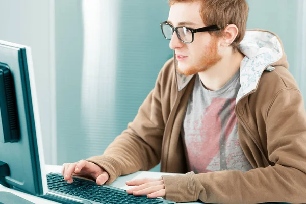 年轻的千年工作者站在电脑屏幕前 戴着黑色的处方药眼镜 棕色的帽衫 他在打字 非常有兴趣和专注 — 图库照片