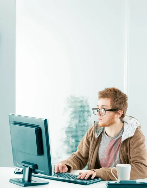 宽射门与宽铜板 年轻的书呆子千年工作者站在电脑屏幕前 戴着黑色的处方药眼镜 棕色的帽衫 他在打字 非常有兴趣和专注 — 图库照片