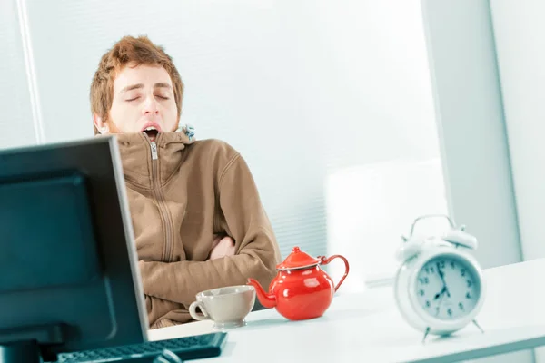 荷兰人拍摄了一个寒冷和困倦 颤抖的年轻工人打呵欠时 他蜷缩在他的棕色运动衫 他在办公室里吃早餐 桌上有一个杯子和一个茶壶 放在电脑旁边 — 图库照片