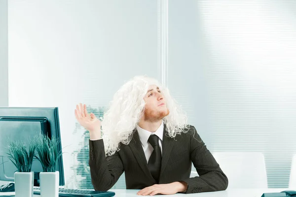 Young man goofs around the office in a white long-haired woman's wig, perhaps even a white beard used as a wig. He struts around like a show business diva, raising a hand to pose with arrogance. Humor