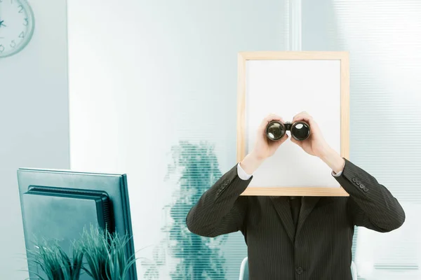 コンピュータモニタも見ることができるオフィスで囲まれたサインヘッドを持つ男 男は立ち上がって双眼鏡であなたを見ている ホワイトスペースは編集無料 — ストック写真