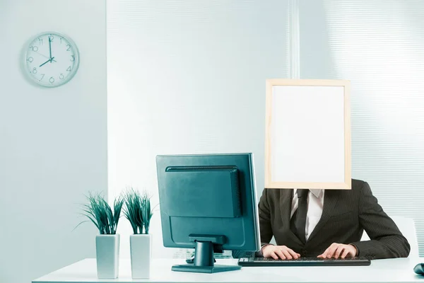 Man with framed sign head in an office works on computer. Visible body and arms work at keyboard and monitor can be seen. The head is available for graphic processing.
