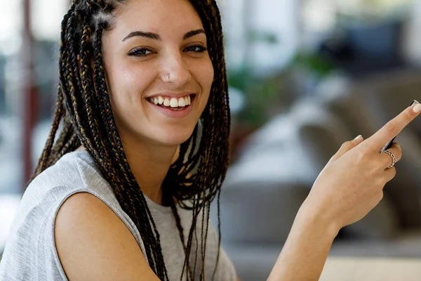 Candid close-up portrait of a young woman laughing while using a cell phone, blurred background. She has a nose piercing, big teeth, box braids, groomed eyebrows, possibly freckles, and looks stunning
