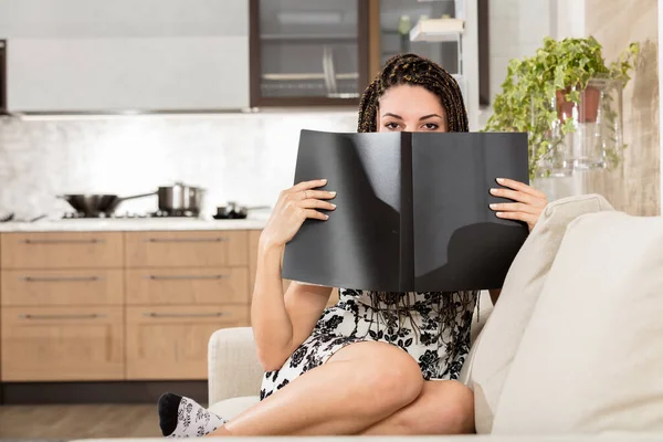 年轻的女人坐在客厅的沙发上 后面有一个现代化的厨房 她把脸藏在一本黑色的封面小册子或笔记本后面 平静而迷人的凝视着外面 她的头发是盒状的 — 图库照片