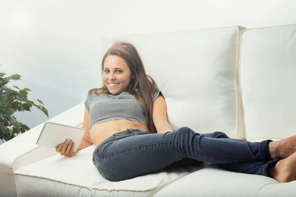 ソファに座っている若い女性は デジタルタブレットを使用して 長い足でリラックスしたジーンズとトーンのAbs樹脂製のTシャツを着て 足を伸ばして裸足でリラックスしました 彼女は若くて美しく聡明だ — ストック写真