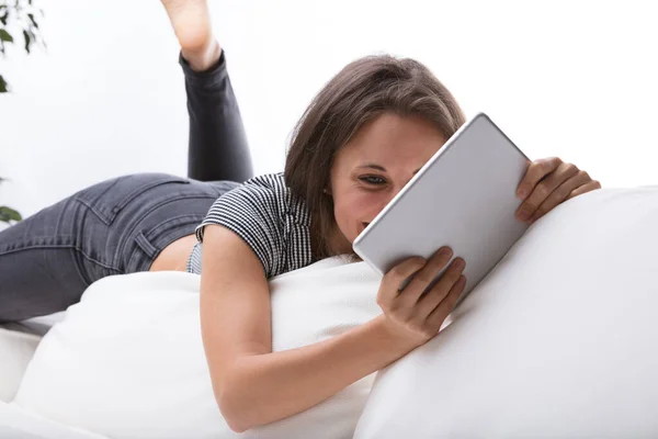 年轻女人躺在白色沙发靠背的边缘上 因为身体合适而懒洋洋地扭曲着身体 看着数字平板电脑上看不见的屏幕上的什么东西 也许是视频 书籍或电脑 — 图库照片