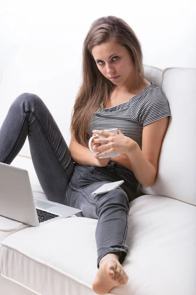 若い女性がノートパソコンの前でマグカップから暖かいものを飲んでいるソファに座っている 彼女は裸足でリラックスしている 彼女の視線は挑戦的で深刻であります まだ魅力的でセクシー 彼女はセルだ — ストック写真