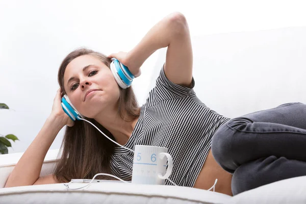 女孩侧卧在沙发上 双腿蜷缩在床上 用手机耳机听音乐 用手拿着耳机 一边喝着热的东西 所有的东西都是白的 — 图库照片