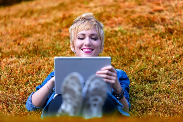Frontal long shot portrait of a young woman lying on the side of a hill, smiling while using a digital tablet, surrounded by the colors of nature, on the grass of a meadow, with short blonde hair and