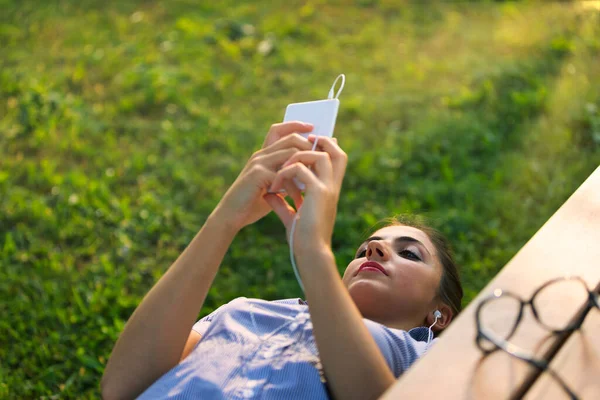 上の3分の1のビューでは ベンチに横になっている女の子が 画面に集中しているときにスマートフォンに接続されている有線イヤフォンを示しています 彼女はビデオ通話やEnjに従事しているかもしれません — ストック写真