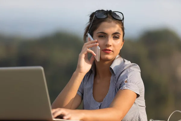 一位年轻 专心致志的女性 在高速互联网的帮助下 在一个美丽的日子里 正在从事组织和沟通工作 在电话上交谈 并在室外使用笔记本电脑 她是认真的 — 图库照片