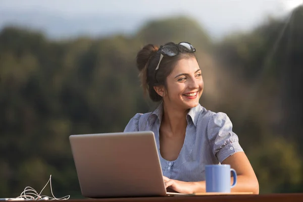 A young woman working on her tablet outdoors on a sunny day stops and smiles at something or someone to her right. She wears sunglasses like a headband and a sunbeam enters the frame. Mountains are in