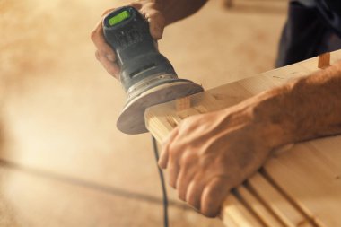 The carpenter's hands grip a sander. The wood artisan, an experienced professional, holds a board or piece of wood with strong and precise hands, carefully sanding it with a machine that vibrates sand clipart