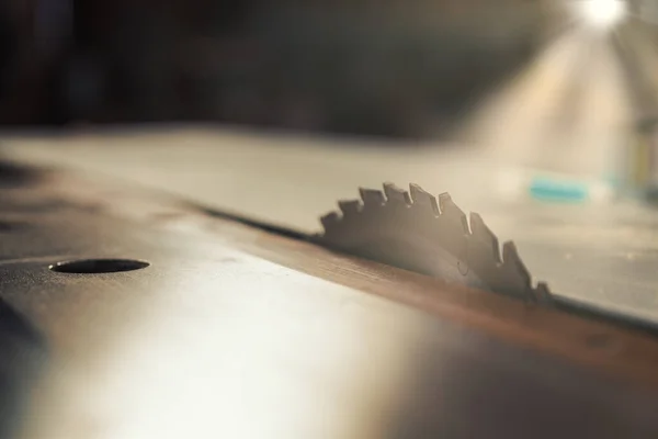 Rays of light penetrating through the large window of a woodworking shop strike the toothed blade of a modern wood saw. The contemporary craftsman is a technological woodworker.
