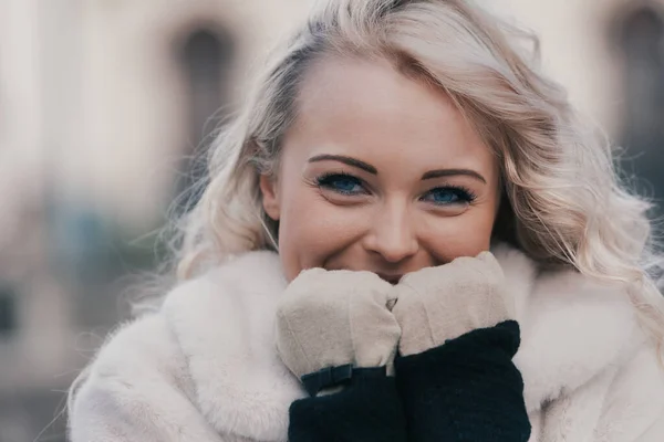 Frontal portrait of a blond woman with blue eyes clutching the warmth of her gloves her smiling face. She has a fur coat and long hair with ringlets.