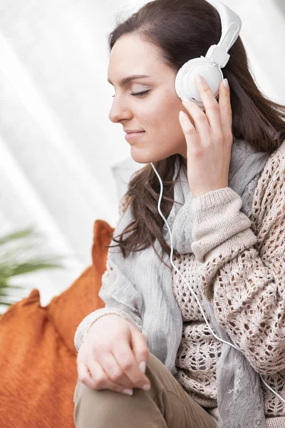 一个穿着褐色衣服的女人正赤脚坐在她的沙发上 她在听着带着白色耳机的音乐 或者听着收音机或者一个有趣的播客 放松和娱乐H — 图库照片