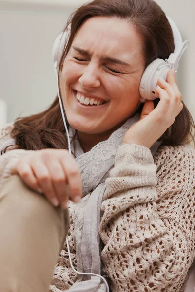 一个穿着褐色衣服的女人正赤脚坐在她的沙发上 她在听着带着白色耳机的音乐 或者听着收音机或者一个有趣的播客 放松和娱乐H — 图库照片
