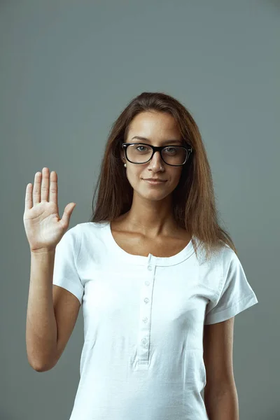 Frontal image of a young woman, glasses, white shirt, hand up, five fingers raised, depicting number five, greeting, one-through-five series, fifth fraction or hand-raised vote