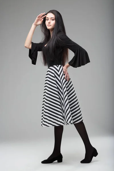 Long-haired young woman modeling in white dress with wide sleeves, herringbone-patterned zebra-striped black and white skirt, black stockings, low-heeled shoes. Elegant, sophisticated, beautiful. Full